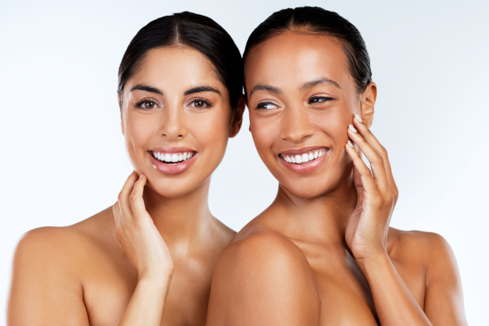 Two smiling women with glowing skin posing together, each gently touching their face, wearing white strapless tops against a light background, showcasing healthy and radiant complexions.