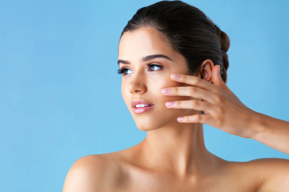 exploring dermatology specialties Woman with clear, glowing skin touching her face gently while looking to the side, posed against a bright blue background — representing healthy skin and dermatology care.